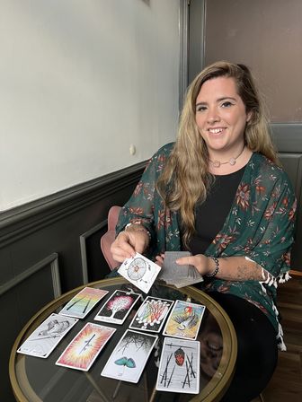 Smiling person in a floral kimono doing a tarot card reading at a small round glass table, holding a card above an array of illustrated tarot cards in a cozy indoor corner.