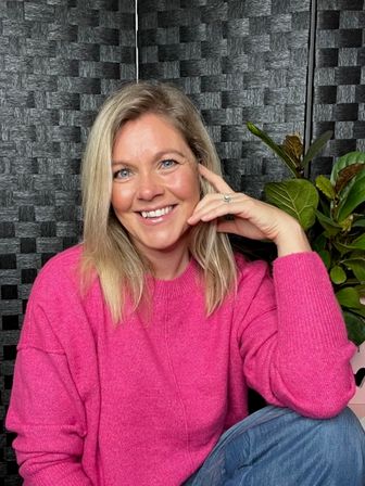 Smiling blonde woman in a bright pink sweater and jeans seated indoors against a textured black panel and green potted plant, casual friendly portrait/headshot.