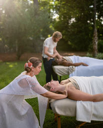 Sunlit garden massage scene on a grassy lawn — two therapists giving outdoor spa treatments on portable tables, a woman with a floral hairpiece stretching a client’s neck while another therapist works on a second client wrapped in white towels.