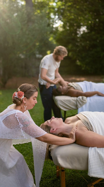 Sunlit garden massage scene on a grassy lawn — two therapists giving outdoor spa treatments on portable tables, a woman with a floral hairpiece stretching a client’s neck while another therapist works on a second client wrapped in white towels.