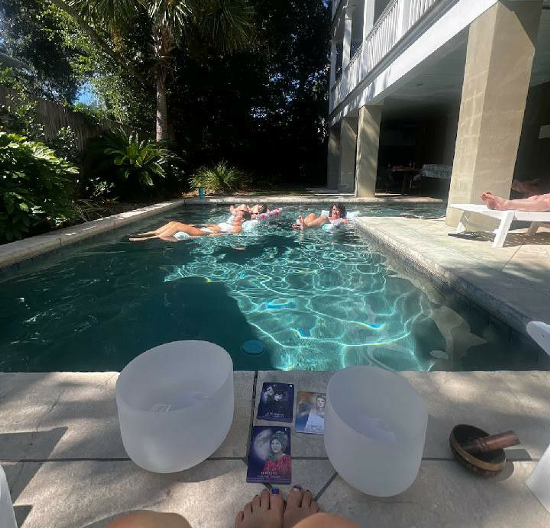 Sunlit backyard pool scene with people floating on inflatables under a shaded patio, palm trees and lush landscaping; crystal singing bowls, oracle cards and bare feet on the pool deck.