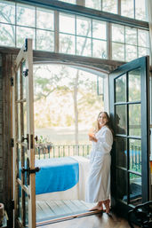Barefoot woman in a white robe standing in open French doors onto a sunlit balcony, holding a glass of rosé and smiling beside a blue-covered daybed with trees outside.