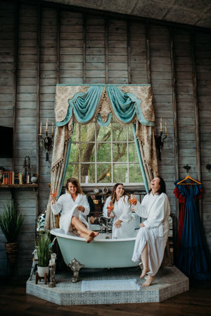 Three women in white robes laughing and toasting rosé from a vintage clawfoot bathtub beneath teal velvet drapery in a rustic wood‑paneled room with a large window and garden view.