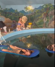 Two people lounging on blue float mats in an indoor pool beneath a colorful tropical mural with palm trees, while a person at the pool edge adjusts a sun hat.