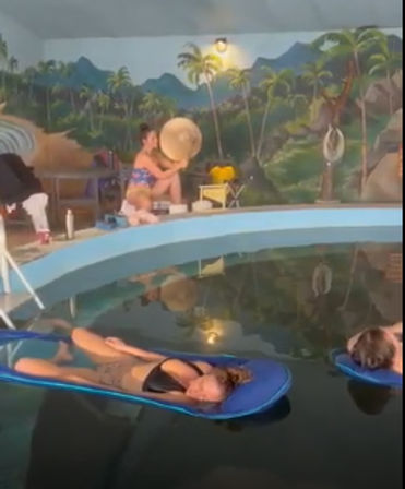Two people lounging on blue float mats in an indoor pool beneath a colorful tropical mural with palm trees, while a person at the pool edge adjusts a sun hat.