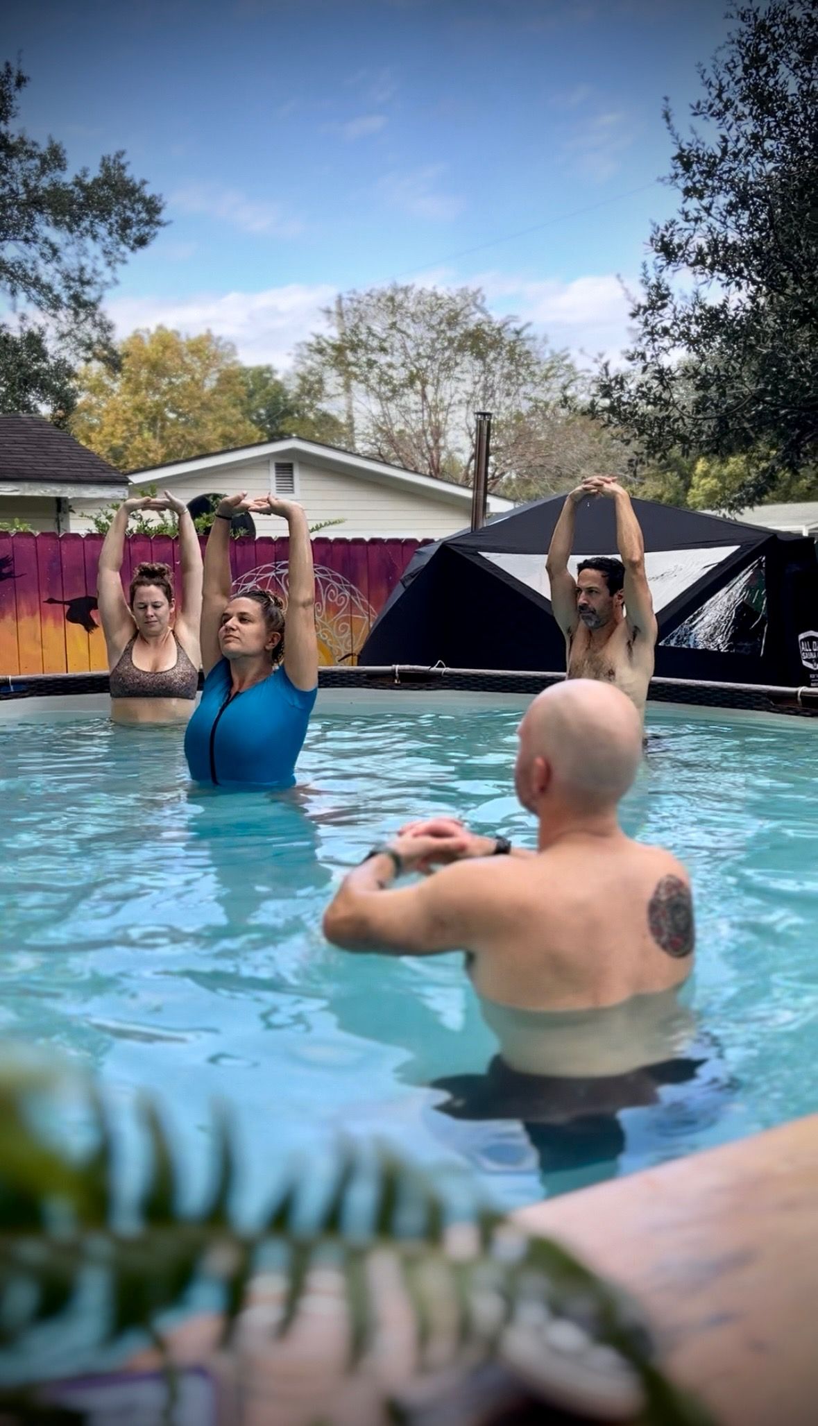 Group of adults doing water yoga stretches in a backyard above-ground pool, arms raised toward a blue sky with a colorful fence and trees in the background.