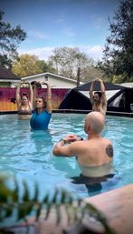 Group of adults doing water yoga stretches in a backyard above-ground pool, arms raised toward a blue sky with a colorful fence and trees in the background.