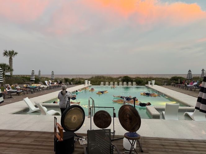 Beachfront pool at sunset with pink clouds over the ocean horizon; people floating on mats in the lit pool while a musician plays gongs on the wooden deck, striped umbrellas and lounge chairs lining the sides.