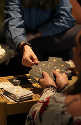 Close-up of a tarot reading: hands exchanging celestial moon-and-star cards over a woven table with stacked decks and crystals, cozy indoor vibe.
