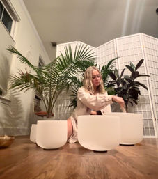 Person playing white crystal singing bowls for a home sound bath, seated on hardwood floor surrounded by large tropical plants and a white folding room divider — cozy meditation wellness scene