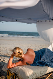 Person lounging on a portable massage table under a beach canopy, wrapped in a blue towel with sandy shore and ocean waves in the background.