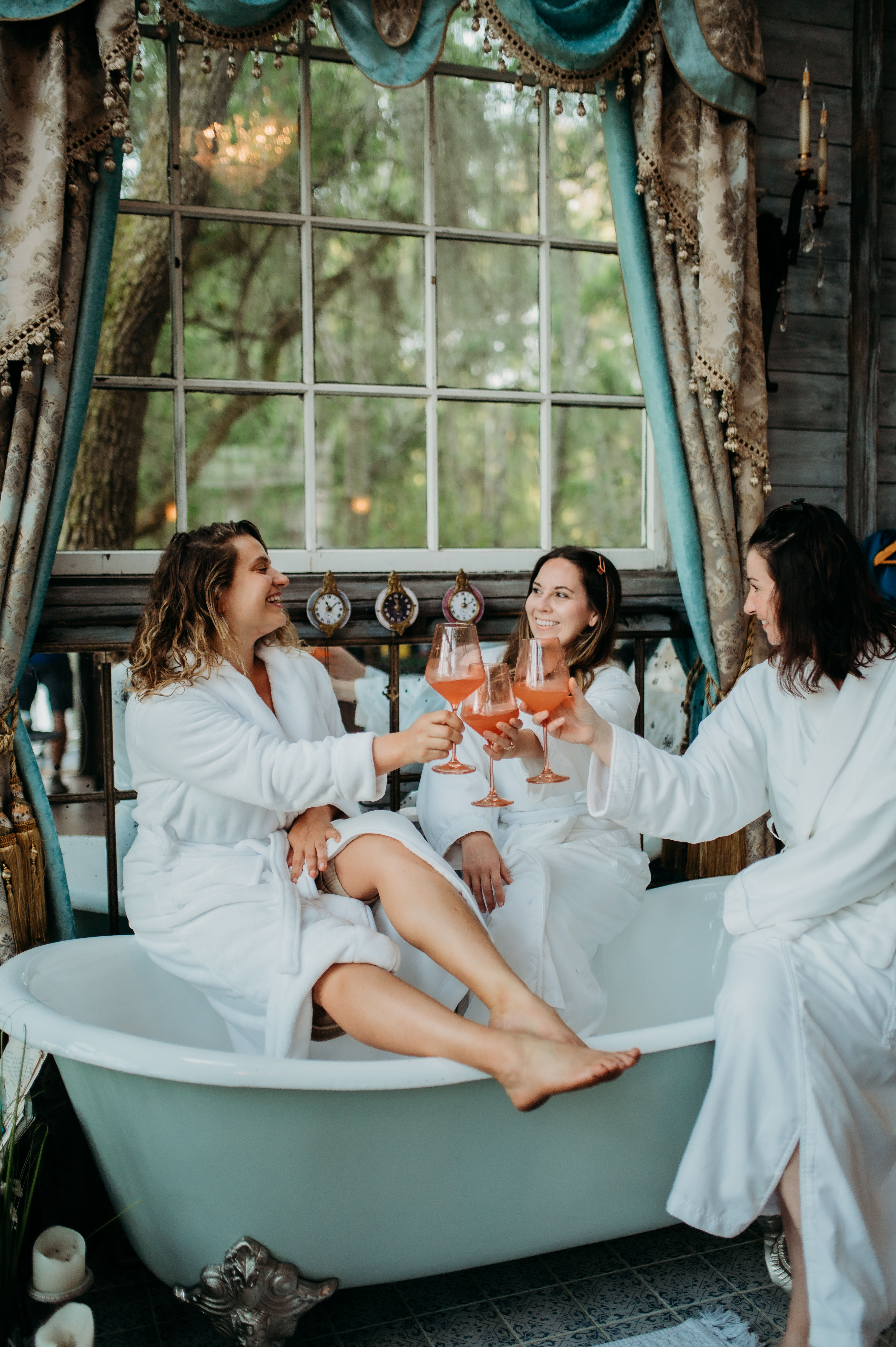 Three women in white robes clinking pink cocktails while lounging in a vintage clawfoot bathtub by a large window with draped curtains and a leafy outdoor view — cozy spa-day girls’ getaway.