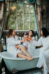 Three women in white robes clinking pink cocktails while lounging in a vintage clawfoot bathtub by a large window with draped curtains and a leafy outdoor view — cozy spa-day girls’ getaway.