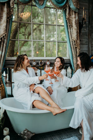 Three women in white robes clinking pink cocktails while lounging in a vintage clawfoot bathtub by a large window with draped curtains and a leafy outdoor view — cozy spa-day girls’ getaway.