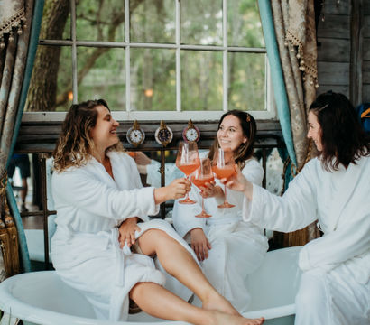 Three women in white robes clinking pink cocktails while lounging in a vintage clawfoot bathtub by a large window with draped curtains and a leafy outdoor view — cozy spa-day girls’ getaway.
