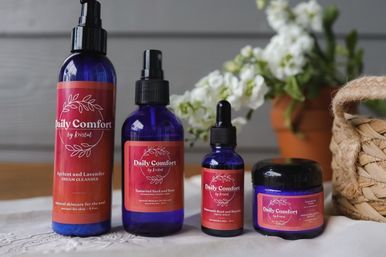 Four cobalt glass skincare bottles and a jar with red labels arranged on a wooden tabletop, white flowers and a woven basket in the background in a home-spa setting.