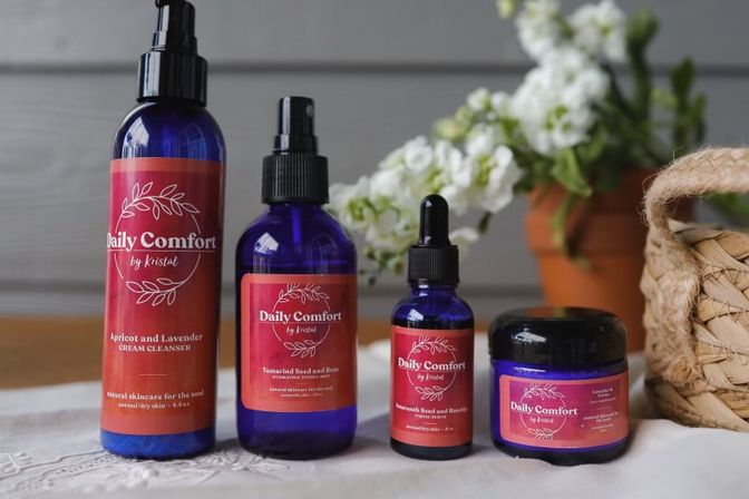Four cobalt glass skincare bottles and a jar with red labels arranged on a wooden tabletop, white flowers and a woven basket in the background in a home-spa setting.