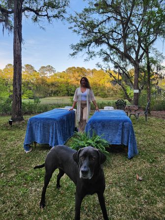 Black dog in foreground with two outdoor massage tables draped in blue, practitioner standing between them beneath oak trees draped in Spanish moss in a peaceful meadow
