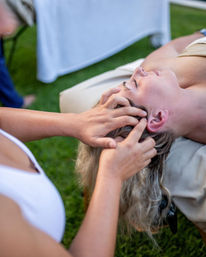 Person lying on an outdoor massage table receiving a relaxing scalp and head massage on a green lawn, wellness spa vibe
