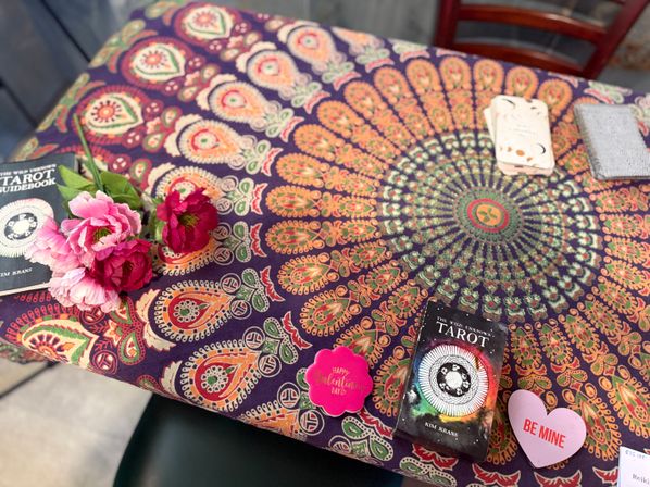 Colorful bohemian mandala tablecloth with a tarot deck and cards, pink faux flowers, and Valentine heart tokens reading "Be Mine".
