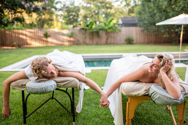 Couple enjoying an outdoor couples massage on portable massage tables beside a backyard pool, holding hands across the tables on a sunny lawn.