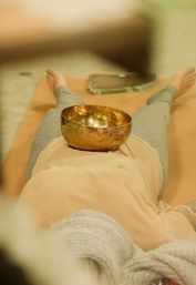 Person lying on a mat with a brass singing bowl on their abdomen during a sound-bath meditation in a cozy wellness space