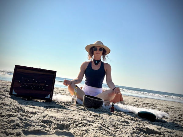 Person meditating cross-legged on a sunny ocean beach wearing a sun hat and sunglasses, surrounded by a singing bowl, small bottles and a wooden box — coastal beach meditation and sound-healing scene.