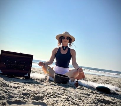 Person meditating cross-legged on a sunny ocean beach wearing a sun hat and sunglasses, surrounded by a singing bowl, small bottles and a wooden box — coastal beach meditation and sound-healing scene.