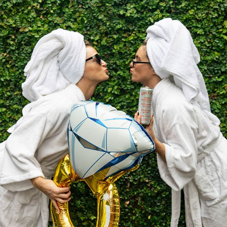 Two people in white bathrobes and towel turbans, wearing sunglasses, leaning in for a playful kiss while holding an inflatable diamond ring balloon and a canned drink in front of a lush green hedge.