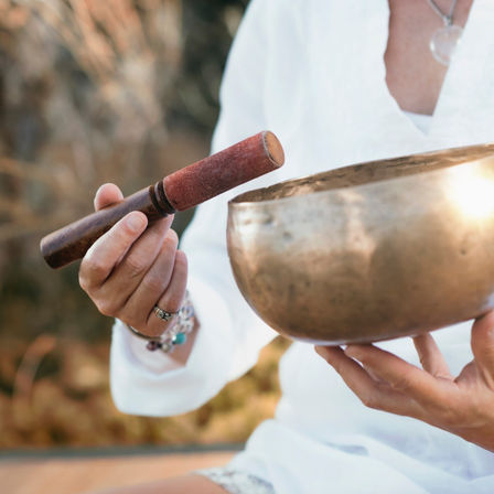 Outdoor meditation scene with hands holding a suede-covered wooden mallet and a bronze singing bowl for sound healing and relaxation.