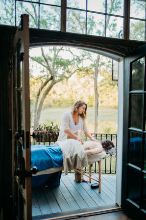 Massage therapist giving a relaxing outdoor back massage to a client on a portable table on a sunlit wooden porch balcony overlooking oak trees and a meadow