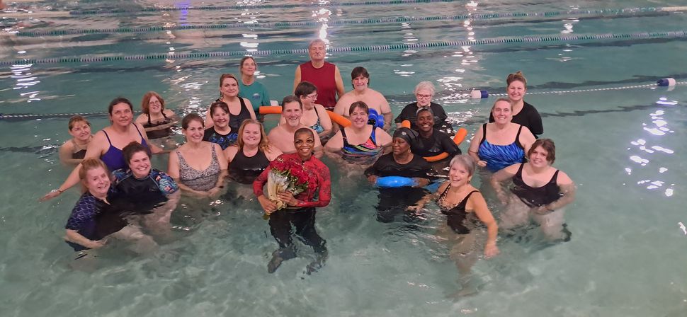 Laughing group of adult swimmers gathered in an indoor community pool, many wearing swimsuits and using pool noodles, one person front-center holding a bouquet of red flowers.