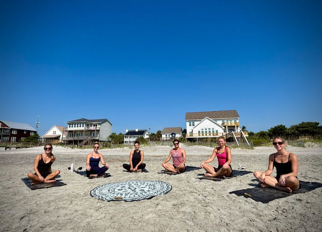 Six people seated for beach yoga on a sandy shore around a round patterned beach towel, with coastal houses behind them and a clear blue sky