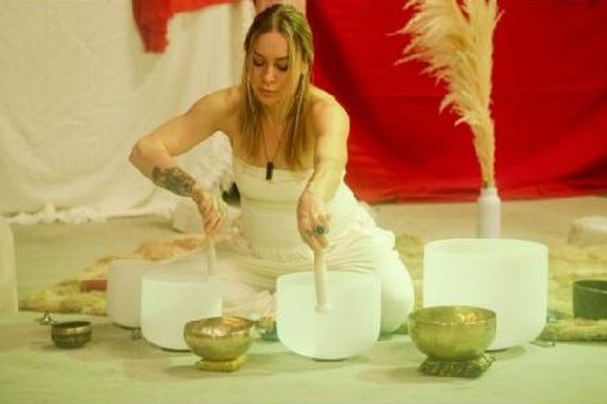 Person in white seated on the floor playing crystal singing bowls and brass bowls with mallets during a sound-bath session in a softly lit indoor space with red backdrop and pampas grass.