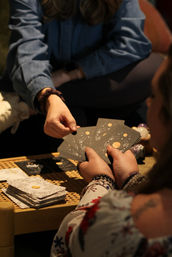 Close-up of hands exchanging celestial tarot cards over a woven table with a stack of cards and crystals during a cozy indoor tarot reading, beaded bracelets visible.