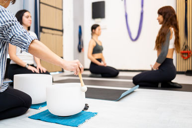 Women seated on yoga mats in a bright wellness studio for guided meditation and sound bath, instructor striking a white crystal singing bowl.