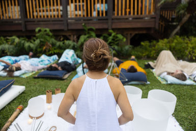 Person in a white top leading an outdoor sound bath on a grassy garden lawn with crystal singing bowls and participants relaxing on mats and blankets
