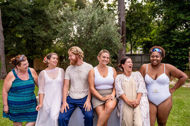Six adults sitting together on a bench in a sunny backyard — laughing friends in summer dresses, casual clothes and a white swimsuit with green lawn and trees in the background, outdoor summer gathering.