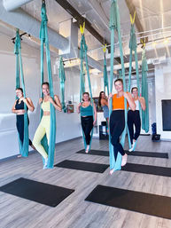 Group of six women smiling in an aerial yoga class suspended in teal fabric hammocks above black mats in a bright modern fitness studio with light wood floors.