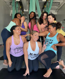Eight women in colorful tank tops laughing and posing in an indoor aerial yoga studio with teal silks and yoga mats, group fitness vibe.