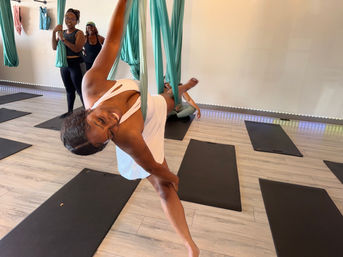 Smiling woman suspended sideways in a teal aerial yoga hammock during an indoor aerial yoga class, striking a balance pose while others watch, black mats on light wood floor