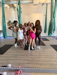 Six women in colorful activewear taking a mirror selfie in an aerial yoga studio with teal silk hammocks, yoga mats and light wood floors.