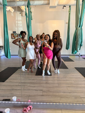 Six women in colorful activewear taking a mirror selfie in an aerial yoga studio with teal silk hammocks, yoga mats and light wood floors.