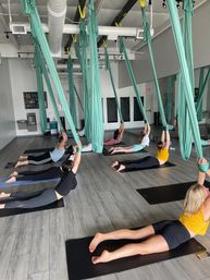 Group aerial yoga class in a bright fitness studio with participants lying on mats reaching up to teal aerial hammocks suspended from the ceiling