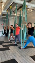 Smiling women in a bright aerial yoga class using teal silk hammocks in a modern fitness studio with wood floors and black mats.