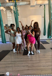 Mirror selfie of six women in colorful activewear smiling and posing in an aerial yoga studio with teal hammocks, yoga mats on light wood floors, and exposed ceiling ductwork — group fitness/aerial yoga class vibe.