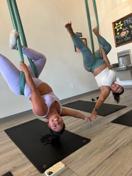 Two women smiling upside-down in aqua aerial yoga hammocks, holding hands above black mats in a bright indoor aerial yoga studio — playful partner fitness pose.