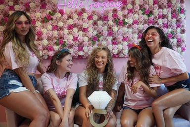 Five friends in matching pink shirts and denim shorts laughing on a couch in front of a pink rose wall and “Hello Gorgeous” neon sign, holding a giant faux diamond ring — playful bachelorette party photo op.