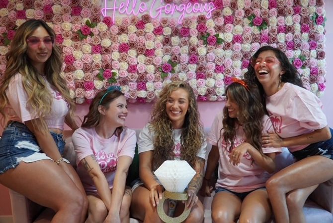 Five friends in matching pink shirts and denim shorts laughing on a couch in front of a pink rose wall and “Hello Gorgeous” neon sign, holding a giant faux diamond ring — playful bachelorette party photo op.