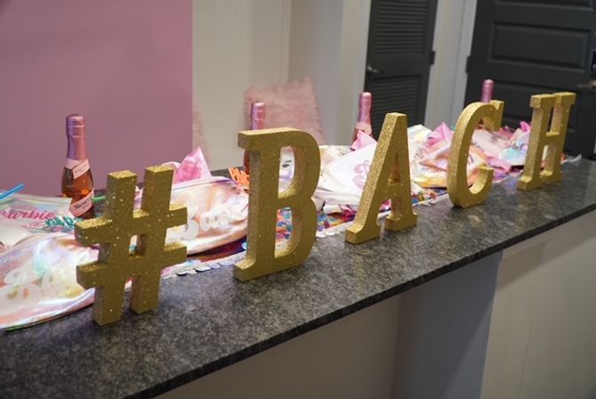 Gold glitter letters spelling "#BACH" on a granite countertop with pink mini champagne bottles and pastel bachelorette party favor pouches